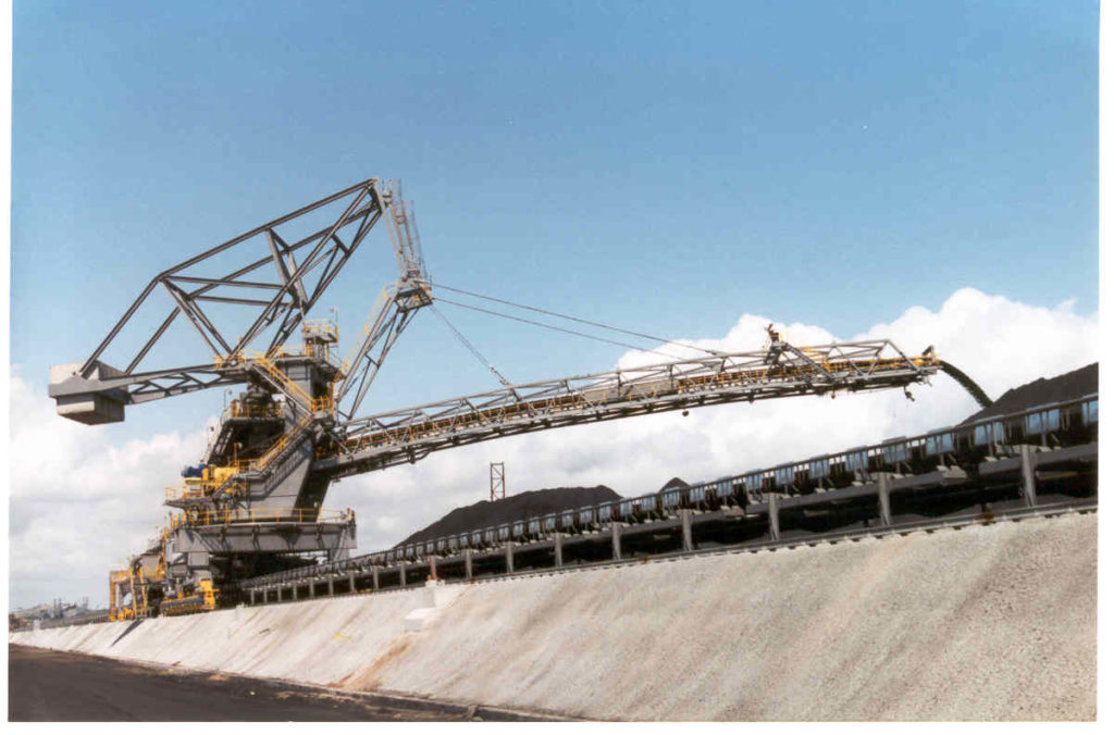 Articulated stacker with long conveyor boom operating alongside a bulk material stockpile.