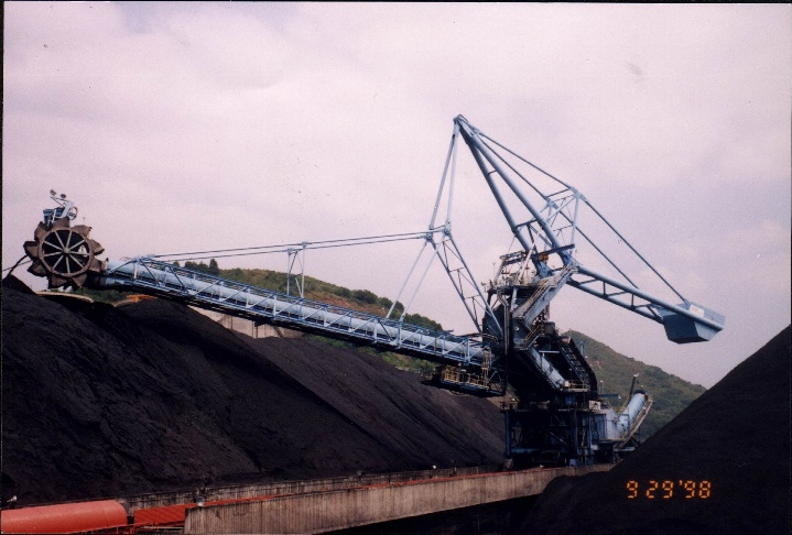 C frame stacker reclaimer operating beside a bulk material stockpile with bucketwheel and conveyor boom visible.
