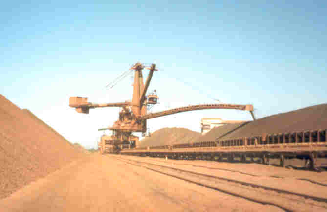 Rail-mounted stacker with a winch-operated luffing boom positioned between bulk material stockpiles.